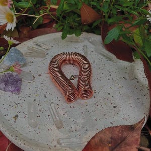 Könnte beinhalten: Ein kupferfarbener Spulenanhänger mit einer kleinen goldenen Perle, der auf einer gesprenkelten, hellfarbigen Platte liegt. Klare Quarzkristalle und ein violetter Amethyst sind um die Platte herum verstreut, mit grünem Laub und Herbstblättern im Hintergrund.