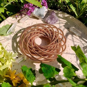 Könnte beinhalten: Eine Sammlung kupferfarbener, verdrehter Metallarmreifen, die spiralförmig auf einer hellen, runden Oberfläche angeordnet sind. Das Bild ist von Grün, Blumen und Kristallen umgeben und schafft eine natürliche, erdige Ästhetik.
