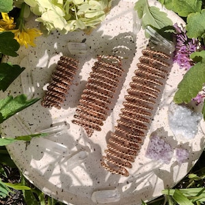 May include: Three copper-coloured wire-wrapped crystal wands of varying sizes rest on a speckled, off-white surface, surrounded by greenery, flowers, and clear quartz crystals. The wands are arranged diagonally.