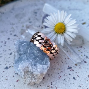 Könnte beinhalten: Ein Kupferring mit einem einzigartigen, strukturierten Design. Der Ring besteht aus kleinen, überlappenden Metallteilen, die ein schuppiges oder kettenartiges Muster erzeugen. Der Ring liegt auf einem Stück blauen Kristall.