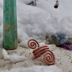 May include: A copper-coloured, spiral-shaped ring sits on a snowy surface beside a tall, green crystal. The ring's design features intricate twists. The background includes snow-covered branches and other crystals.