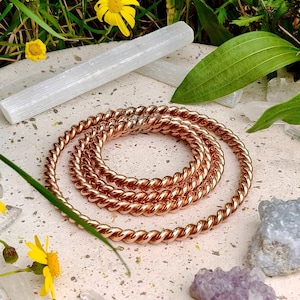 Op de afbeelding: Een opgerolde koperen armband met een gedraaid touwontwerp. De armband wordt getoond op een gespikkelde, lichtgekleurde ondergrond, omringd door kristallen, gele bloemen en groen gebladerte. Er is ook een witte selenietstaaf aanwezig.