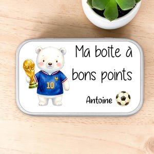 Peut inclure: Une boîte métallique rectangulaire illustrée d'un ours blanc portant un maillot de football bleu avec le numéro 10 et tenant un trophée doré. Le texte indique Ma boîte à bons points et Antoine, avec un ballon de football noir et blanc.