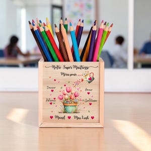 Peut inclure: Un pot à crayons en bois rempli de crayons de couleur. Le pot est décoré d'un motif floral et du texte "Notre Super Maîtresse Merci pour..." avec des noms en dessous.