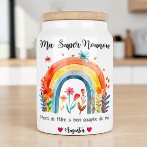 Peut inclure: Un bocal de rangement en céramique blanche avec un couvercle en bois clair, orné d'une illustration aquarelle de fleurs et d'un arc-en-ciel. Le texte indique "Ma Super Nounou", "Merci de t'être si bien occupée de moi" et "Augustin" avec deux cœurs.