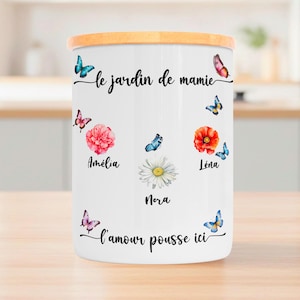 Peut inclure: Un pot en céramique blanc avec un couvercle en bois, décoré de papillons et de fleurs à l'aquarelle. Le pot porte l'inscription "le jardin de mamie" et des noms comme "Amélia" et "Léna". La phrase "l'amour pousse ici" est également écrite sur le pot.
