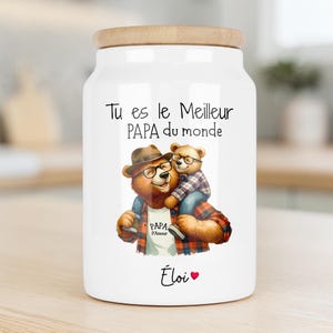 Peut inclure: Un bocal de rangement en céramique blanche avec un couvercle en bois clair, illustré d'un grand ours à lunettes portant un chapeau et un petit ours sur ses épaules. Le texte indique Tu es le Meilleur PAPA du monde, PAPA D'Amour et le prénom Éloi avec un cœur.