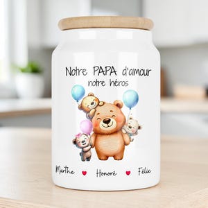 Peut inclure: Un bocal de rangement en céramique blanche avec un couvercle en bois clair, illustré d'un grand ours brun entouré de trois petits oursons et de ballons colorés. Le texte indique "Notre PAPA d'amour notre héros" avec les prénoms "Marthe", "Honoré" et "Félix".