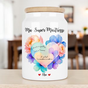 Peut inclure: Un bocal de rangement en céramique blanche avec un couvercle en bois clair. L'avant présente un motif de cœur floral aux couleurs pastel, le texte "Ma Super Maitresse", la phrase "Difficile à quitter, Dure à trouver, et surtout Impossible à oublier" et "Elsa".