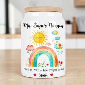 Peut inclure: Un bocal de rangement en céramique blanche avec un couvercle en bois clair, orné d'une illustration aquarelle d'un soleil souriant, d'un arc-en-ciel, de nuages et de fleurs. Le texte indique Ma Super Nounou, Merci de t'être si bien occupée de moi, Célestin.