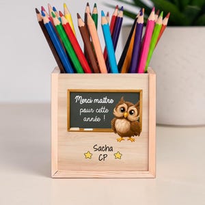 Peut inclure: Un pot à crayons carré en bois rempli de crayons de couleur. La face avant présente un tableau noir illustré d'un hibou avec le texte "Merci maître pour cette année !" ainsi que le nom "Sacha" et "CP" entourés de deux étoiles jaunes.