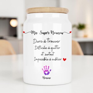 Peut inclure: Pot en céramique blanc avec un couvercle en bois. Le pot porte l'inscription noire "Ma Super Nounou, Dure à trouver, Difficile à quitter et surtout Impossible à oublier." Une empreinte de main violette et le nom "Romane" sont également présents.