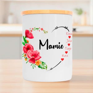 Peut inclure: Un récipient en céramique blanc avec un couvercle en bois. Le récipient est décoré d'un motif floral en forme de cœur rouge et vert, du mot "Mamie" en noir et des noms Elena, Julia, Tiago et Nora.
