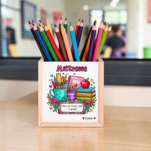 Peut inclure: Un pot à crayons carré en bois rempli de crayons de couleur assortis. La face avant présente un graphique avec le texte "Maîtresse", "Enseigner Aimer Inspirer", "Merci de m'avoir aidé à grandir" et "Ezekiel", entouré d'illustrations de livres, d'une pomme et de fleurs.