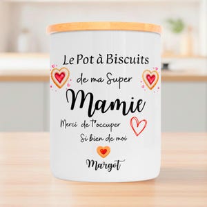 May include: A white ceramic biscuit jar with a wooden lid. The jar is decorated with text and heart-shaped designs. The text includes the words "Le Pot à Biscuits de ma Super Mamie" and "Margot".