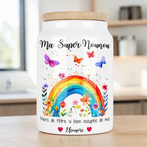 Peut inclure: Un bocal de rangement en céramique blanche avec un couvercle en bois, orné d'un arc-en-ciel, de fleurs et de papillons. Le texte Ma Super Nounou, Merci de t'être si bien occupée de moi, et Eleonore avec deux cœurs rouges est imprimé.