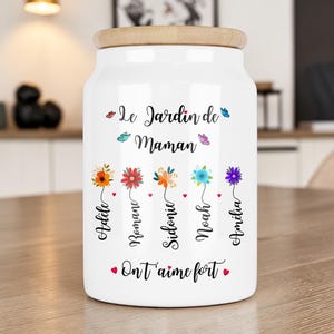 Peut inclure: Un pot en céramique blanc avec un couvercle en bois, décoré de fleurs colorées et du texte "Le Jardin de Maman". Le pot présente des noms et la phrase "On t'aime fort". Papillons et coeurs complètent le design.