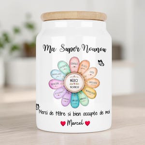Peut inclure: Un bocal de rangement en céramique blanche avec un couvercle en bois clair. L'avant présente un motif de fleur colorée avec des pétales étiquetés en français, entouré du texte "Ma Super Nounou", "Merci de t'être si bien occupée de moi" et "Marcel" avec deux cœurs rouges.