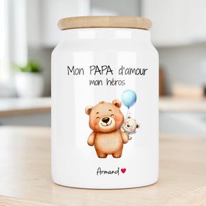 Peut inclure: Un bocal de rangement en céramique blanche avec un couvercle en bois clair. L'avant présente un ours brun tenant un ballon bleu, un petit ours blanc, et le texte "Mon PAPA d'amour mon héros" au-dessus du prénom "Armand" et d'un cœur rouge.