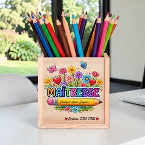 Peut inclure: Un pot à crayons en bois rempli de crayons de couleur. Le pot est décoré d'un motif floral, de papillons et du mot "MAÎTRESSE" avec le texte "Enseigner. Aimer. Inspirer." et le nom "Hortense 2025-2026".