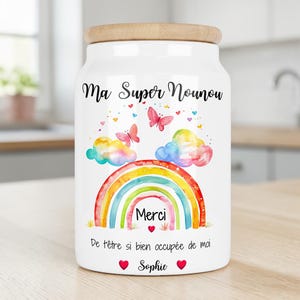 Peut inclure: Un bocal en céramique blanche avec un couvercle en bambou, orné d'un arc-en-ciel aquarelle, de deux papillons roses et de nuages. Le texte indique : Ma Super Nounou, Merci, De t'être si bien occupée de moi, et Sophie, avec des petits cœurs rouges.
