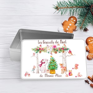 Op de afbeelding: Een rechthoekige, open, zilverkleurige blikken doos met een decoratief deksel. Het deksel heeft een kerstthema-illustratie met een ijsbeer, een hert en andere dieren rond een kerstboom. Tekst op het deksel luidt "Les biscuits de Noël de Mamie Alice."
