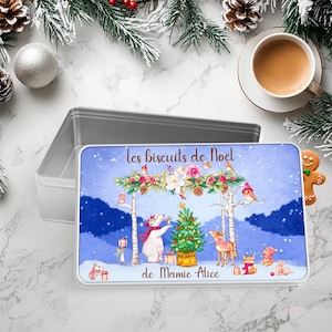 Può includere: Una scatola rettangolare di latta con coperchio, raffigurante una scena invernale con un orso polare, un cervo e altri animali attorno a un albero di Natale. Il testo "Les biscuits de Noël de Mamie Alice" è visualizzato.