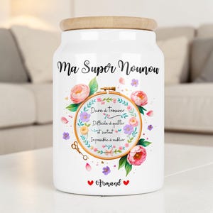 Peut inclure: Un bocal de rangement en céramique blanche avec un couvercle en bois clair, orné du texte "Ma Super Nounou" et d'un motif de tambour à broder floral contenant le texte "Dure à trouver, Difficile à quitter et surtout Impossible à oublier" et le nom "Armand".