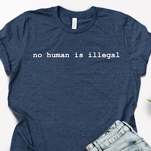 May include: Navy blue t-shirt with the text "no human is illegal" in white. The shirt is paired with light blue denim shorts, sunglasses, and tan sandals. A small succulent plant is also visible.