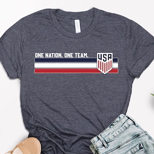 Może przedstawiać: Granatowa koszulka z napisem "ONE NATION. ONE TEAM." i grafiką flagi USA. Koszulka ma czerwono-biało-niebieski wz&oacute;r w paski i logo tarczy USA. Na zdjęciu znajdują się r&oacute;wnież dżinsowe szorty i sandały.