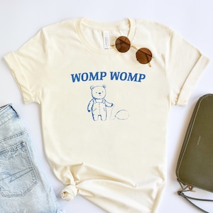 May include: A cream colored t-shirt with short sleeves and a blue graphic of a bear with the text "WOMP WOMP" above it. The t-shirt is laid out on a white surface with a pair of sunglasses, a pair of denim shorts, and a green clutch purse.