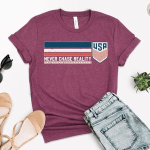 May include: A maroon t-shirt with the text "NEVER CHASE REALITY" and a USA graphic. The graphic includes a blue and red striped design and a shield with the letters "USA". The shirt is paired with denim shorts, sunglasses, and sandals.