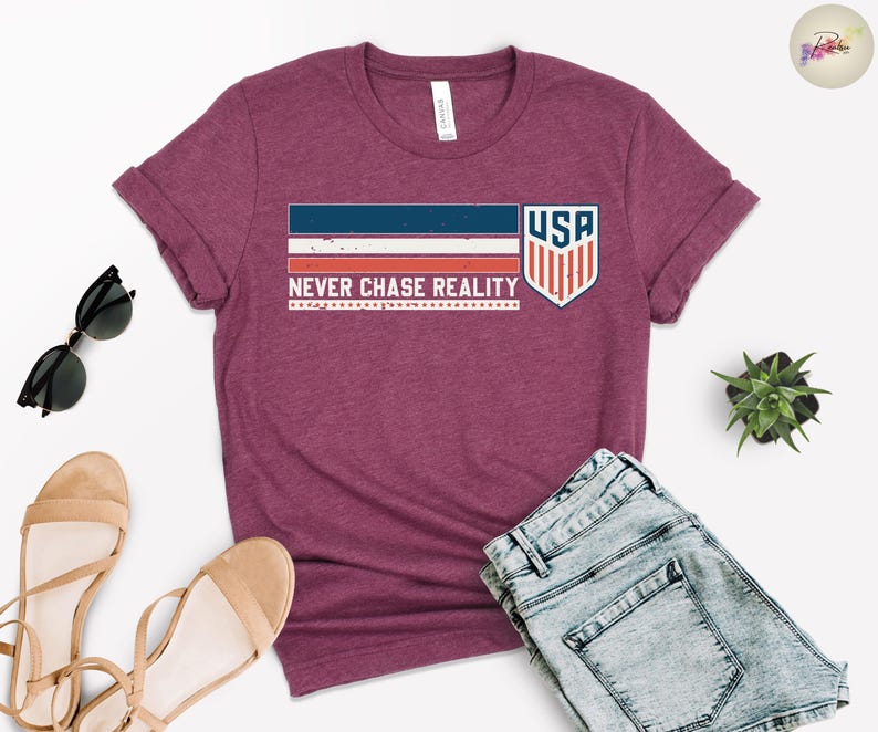 May include: A maroon t-shirt with the text "NEVER CHASE REALITY" and a USA graphic. The graphic includes a blue and red striped design and a shield with the letters "USA". The shirt is paired with denim shorts, sunglasses, and sandals.