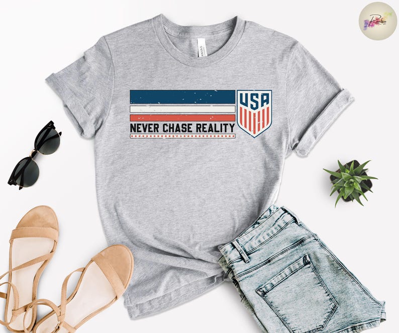 May include: A heather gray t-shirt with a graphic design featuring the text "NEVER CHASE REALITY" and a USA shield logo. The design incorporates red, white, and blue stripes. The shirt is paired with denim shorts, sunglasses, and sandals.