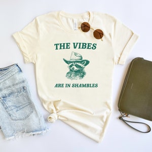 K&ouml;nnte beinhalten: Ein cremefarbenes T-Shirt mit einem gr&uuml;nen Waschb&auml;r, der einen Cowboyhut tr&auml;gt, und dem Text "THE VIBES ARE IN SHAMBLES".