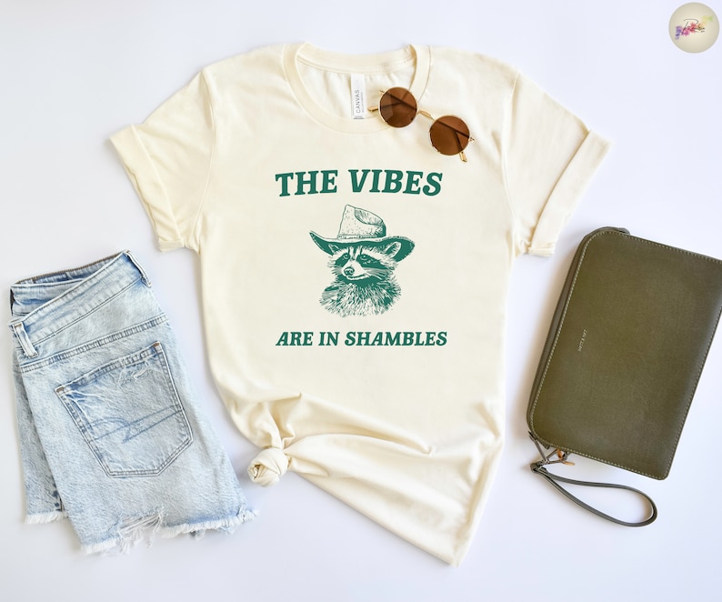 K&ouml;nnte beinhalten: Ein cremefarbenes T-Shirt mit einem gr&uuml;nen Waschb&auml;r, der einen Cowboyhut tr&auml;gt, und dem Text "THE VIBES ARE IN SHAMBLES".