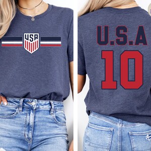 May include: A heather navy blue short-sleeve t-shirt. The front features a small shield graphic with the text USA and horizontal stripes in navy, white, and red. The back displays the text U.S.A. in navy above the large red number 10.