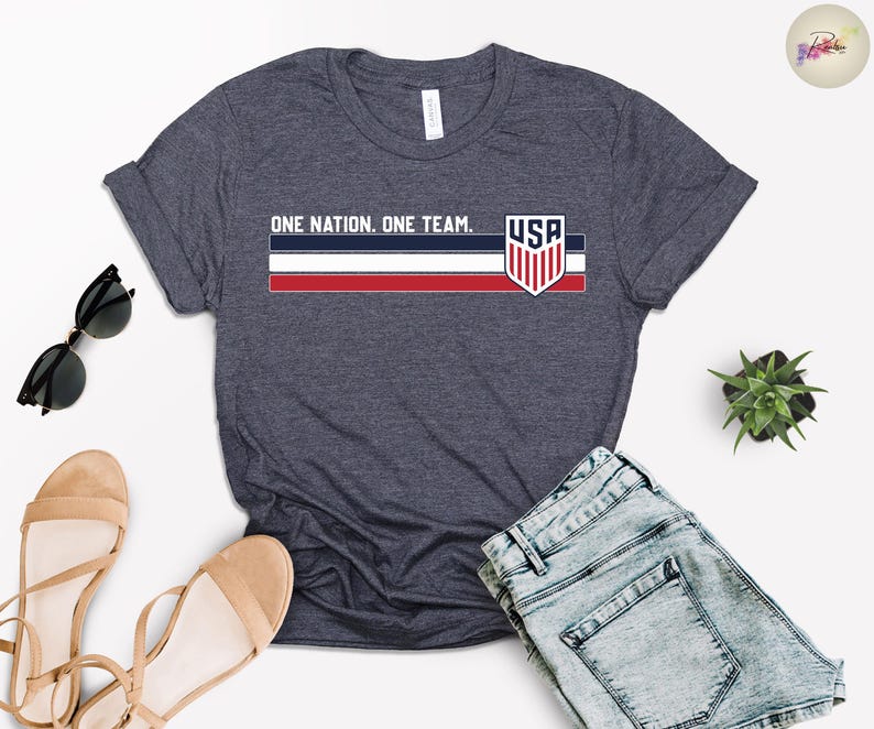 Może przedstawiać: Granatowa koszulka z napisem "ONE NATION. ONE TEAM." i grafiką flagi USA. Koszulka ma czerwono-biało-niebieski wz&oacute;r w paski i logo tarczy USA. Na zdjęciu znajdują się r&oacute;wnież dżinsowe szorty i sandały.