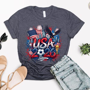 May include: Dark gray t-shirt with a colorful graphic design. The design features the text "USA 2026" with a soccer ball, American flag, eagle, and Statue of Liberty. The shirt is paired with blue denim shorts and sandals.