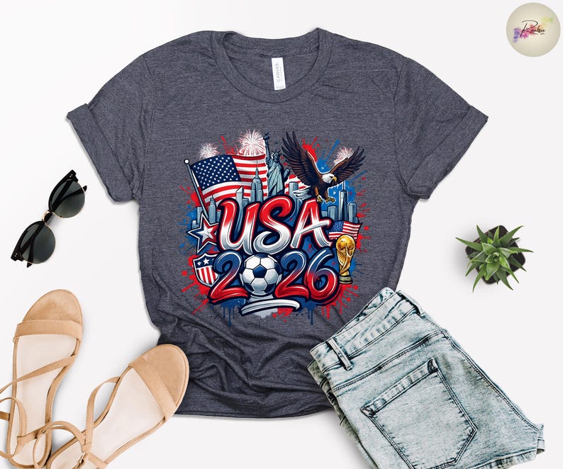 May include: Dark gray t-shirt with a colorful graphic design. The design features the text "USA 2026" with a soccer ball, American flag, eagle, and Statue of Liberty. The shirt is paired with blue denim shorts and sandals.