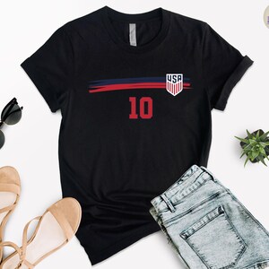 May include: A black short-sleeve t-shirt featuring a horizontal navy blue and red brushstroke graphic across the chest. The design includes a white USA shield emblem on the right side and the large red number 10 centered below the graphic.