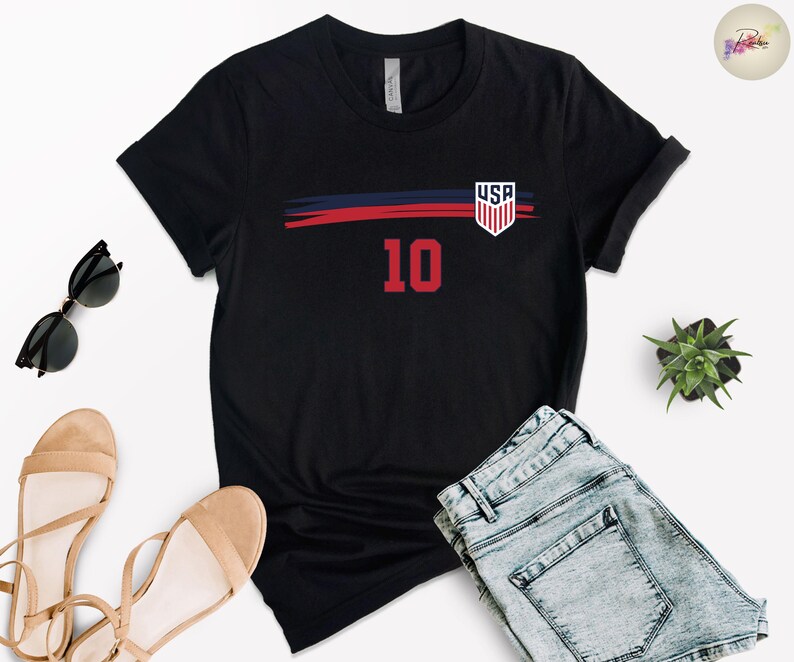 May include: A black short-sleeve t-shirt featuring a horizontal navy blue and red brushstroke graphic across the chest. The design includes a white USA shield emblem on the right side and the large red number 10 centered below the graphic.