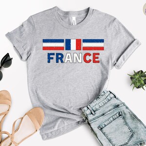 May include: A heather gray t-shirt featuring the word "FRANCE" in blue and red, with a distressed French flag design above. The shirt is paired with denim shorts, sunglasses, and sandals.