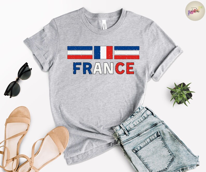 May include: A heather gray t-shirt featuring the word "FRANCE" in blue and red, with a distressed French flag design above. The shirt is paired with denim shorts, sunglasses, and sandals.