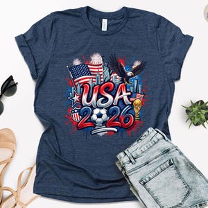 May include: Navy blue t-shirt with a graphic design featuring the text "USA 2026", an American flag, a soccer ball, an eagle, and the Statue of Liberty. The design incorporates red, white, and blue colors.