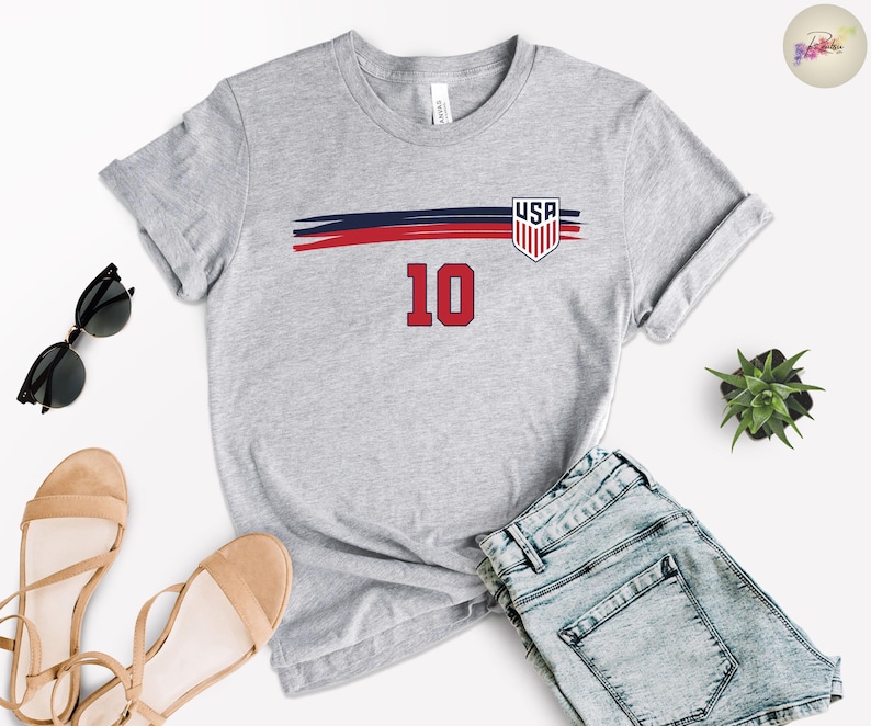 May include: A heather gray short-sleeve t-shirt featuring a horizontal navy and red brushstroke graphic across the chest. The design includes a shield emblem with the text USA and the number 10 in red with a black outline centered below the graphic.