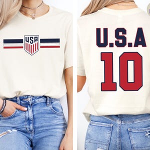 May include: A cream-colored t-shirt featuring a front graphic with a shield containing the text USA, flanked by horizontal navy and red stripes. The back of the shirt displays the text U.S.A. in navy blue above the large red number 10.