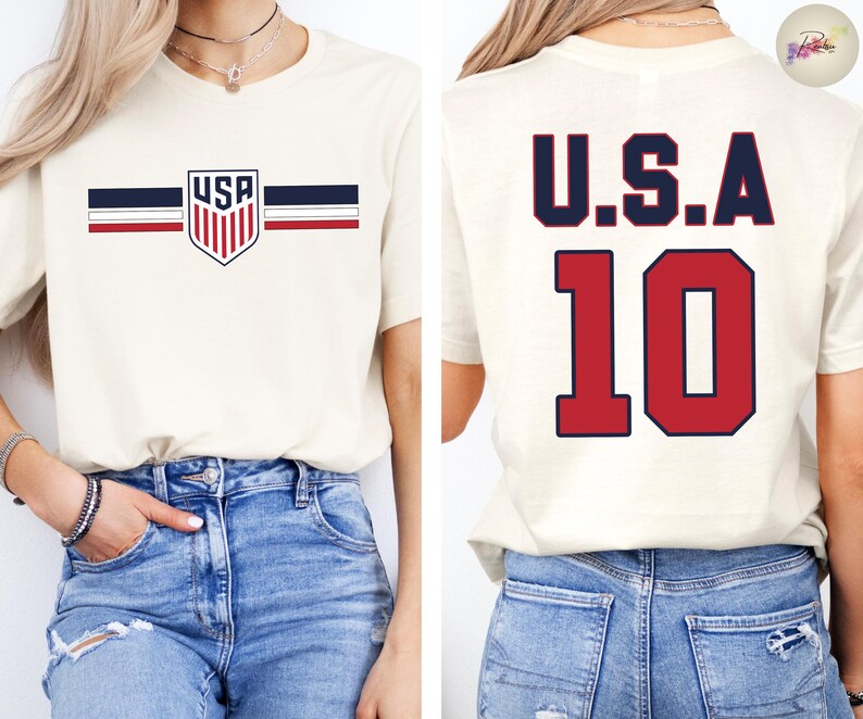 May include: A cream-colored t-shirt featuring a front graphic with a shield containing the text USA, flanked by horizontal navy and red stripes. The back of the shirt displays the text U.S.A. in navy blue above the large red number 10.