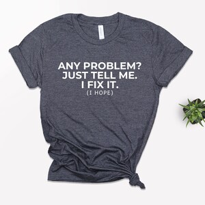 May include: Dark grey t-shirt featuring the white text: "ANY PROBLEM? JUST TELL ME. I FIX IT. (I HOPE)". The shirt is folded and knotted at the bottom. A small potted plant is visible in the background.