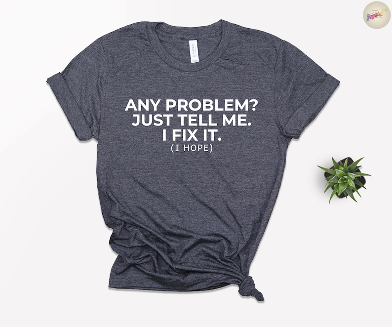 May include: Dark grey t-shirt featuring the white text: "ANY PROBLEM? JUST TELL ME. I FIX IT. (I HOPE)". The shirt is folded and knotted at the bottom. A small potted plant is visible in the background.